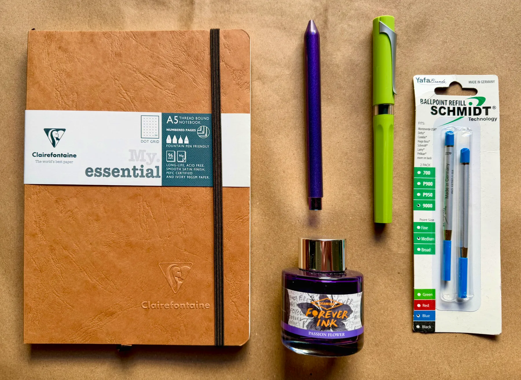 A Clairefontaine journal, Studio Neat Mark One in purple, a green TWSBI Swipe, some Diamine Passion Flower Forever Ink, and a pack of Schmidt EasyFlow 9000 refills arranged on some brown paper.